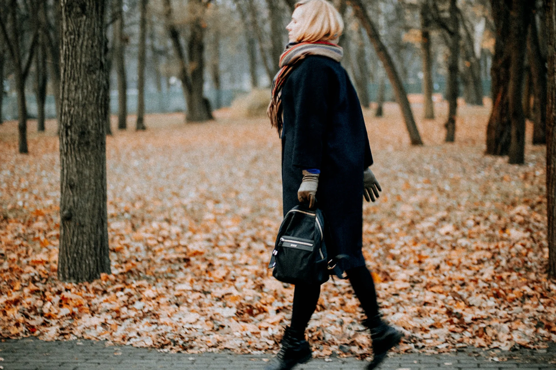 Kobieta w zimowej odzieży idzie przez jesienny park, wśród opadłych liści, z plecakiem.
